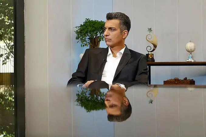 عادل فردوسی&zwnj;پور با کت&zwnj;وشلوار مشکی در استودیوی برنامه فوتبال ۳۶۰ همراه با انعکاس چهره روی میز